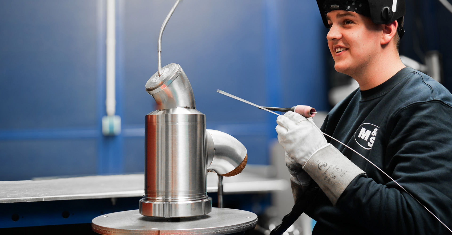 Ein junger Schweißer, der mit einem Lächeln, bei kurzer Schweißpause mit Helmklappe oben, an seinem Schweißdrehtisch sitzt.